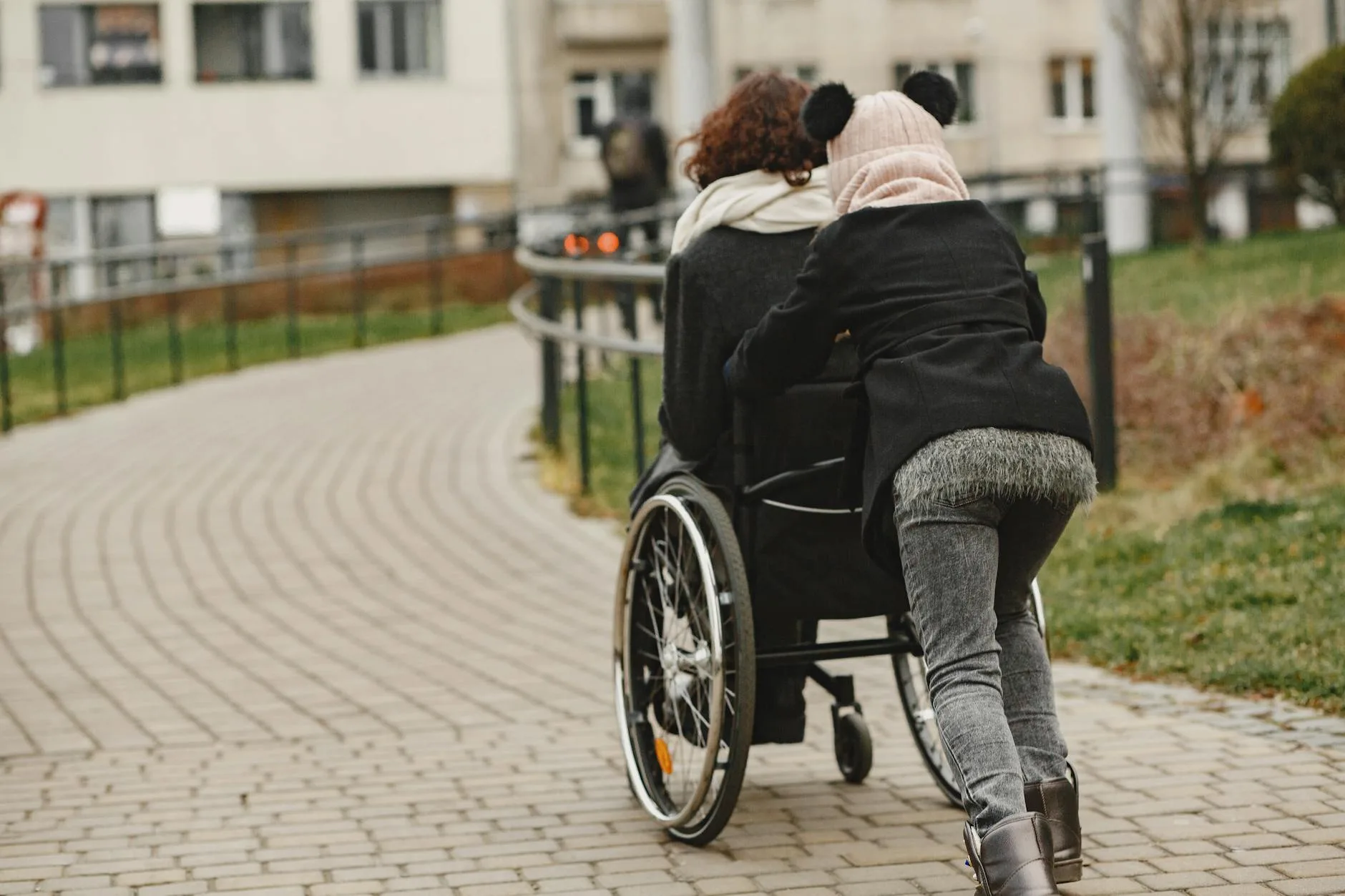 A child supports a woman in a wheelchair during an outdoor walk in a park setting.